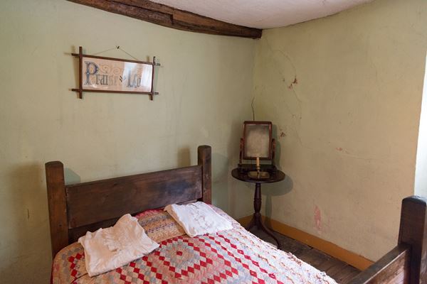 Photo of the main bedroom in the Cottage Museum. There's a wooden double bed covered with a patchwork quilt. A small bedside table stands beside it with a mirror and a candle in a metal holder. Above the head of the bed is an embroidered sampler in a wooden frame, which reads 'Praise the Lord' in elaborate script.. 